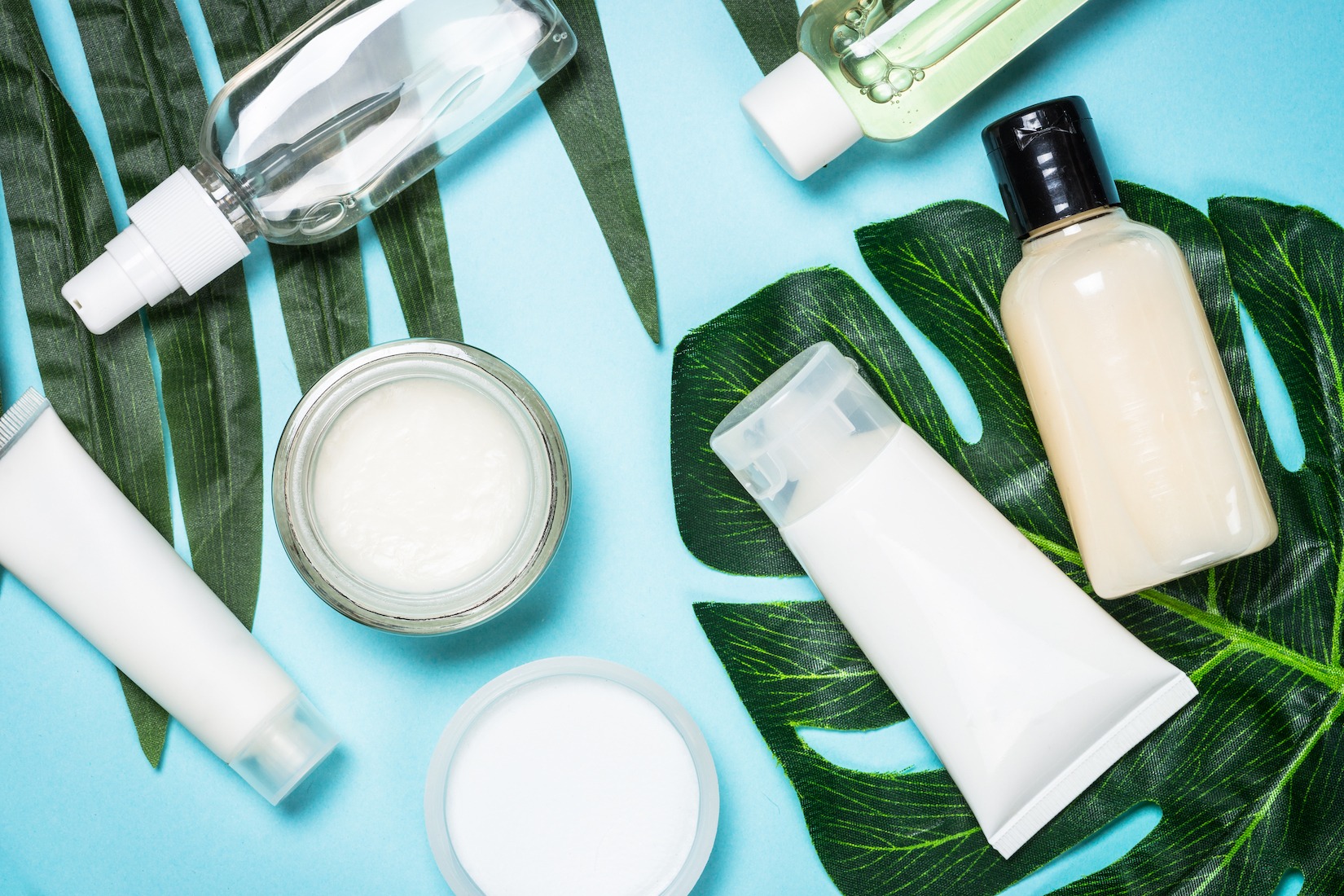 Skin care product, natural cosmetic, cream, tonic, lotion with green leaves. Flat lay image on blue.